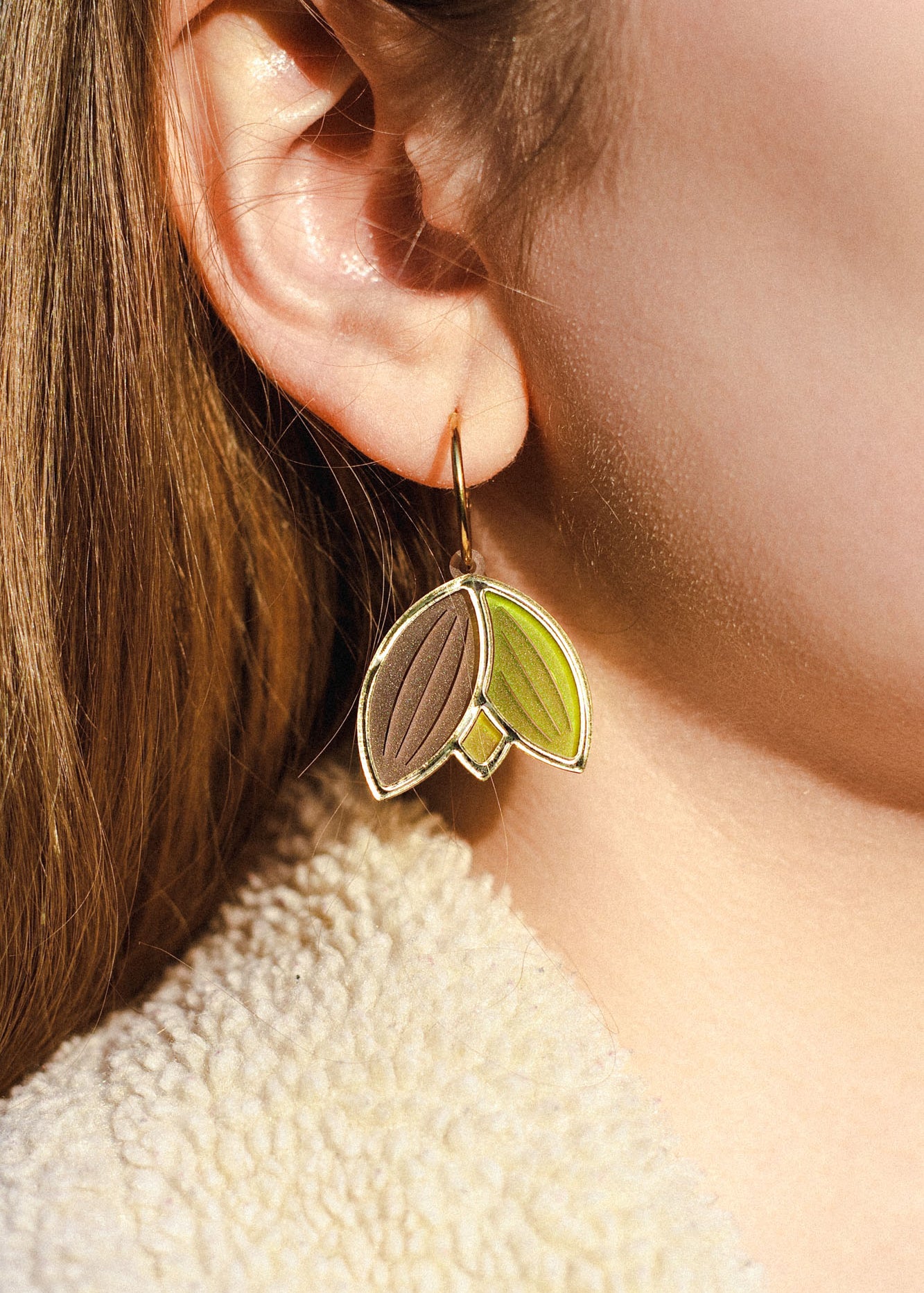 Close-up of a person’s ear wearing handcrafted Tulip Huggie Earrings with three leaf-shaped pendants in brown, green, and tan. The person has light brown hair and is dressed in a cream-colored, textured garment.