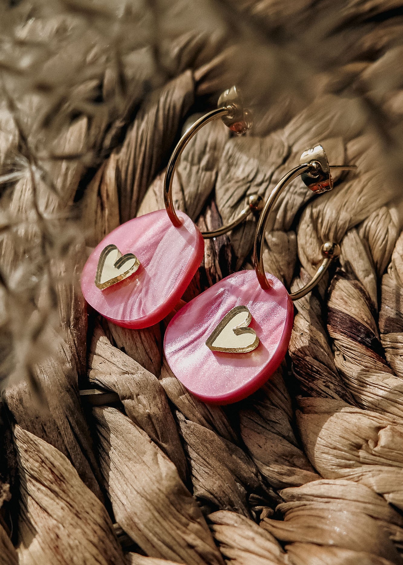 A close-up of lightweight huggie hoops with pink marbled acrylic pendants, each featuring a small gold heart, resting on a woven natural fiber surface—perfect for fans of the Valentine Pebble Earrings.