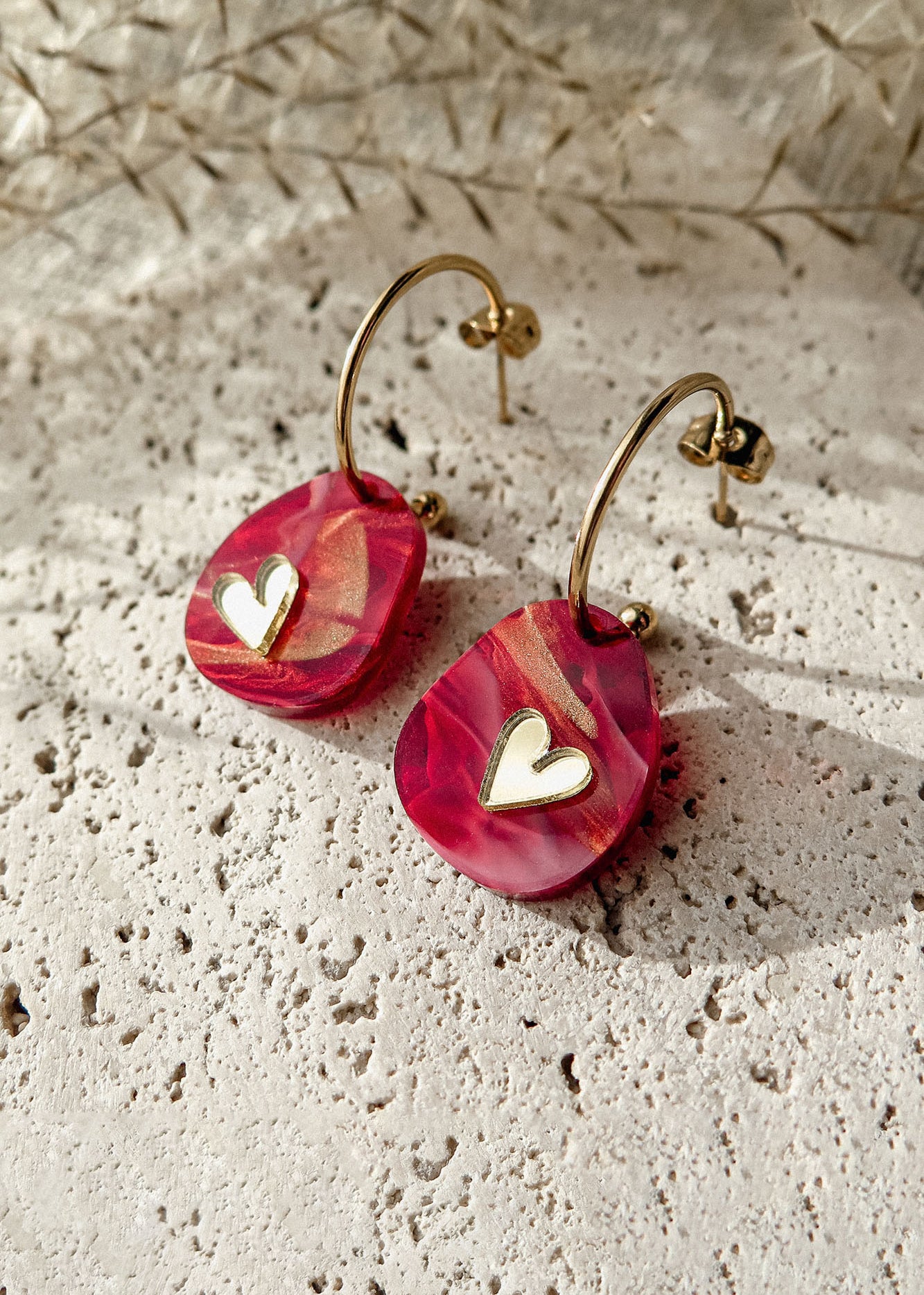 A pair of gold hoop earrings with pink, marbled resin drops and small white heart charms, the Valentine Pebble Earrings are a romantic accessory displayed on a textured stone surface under natural light.