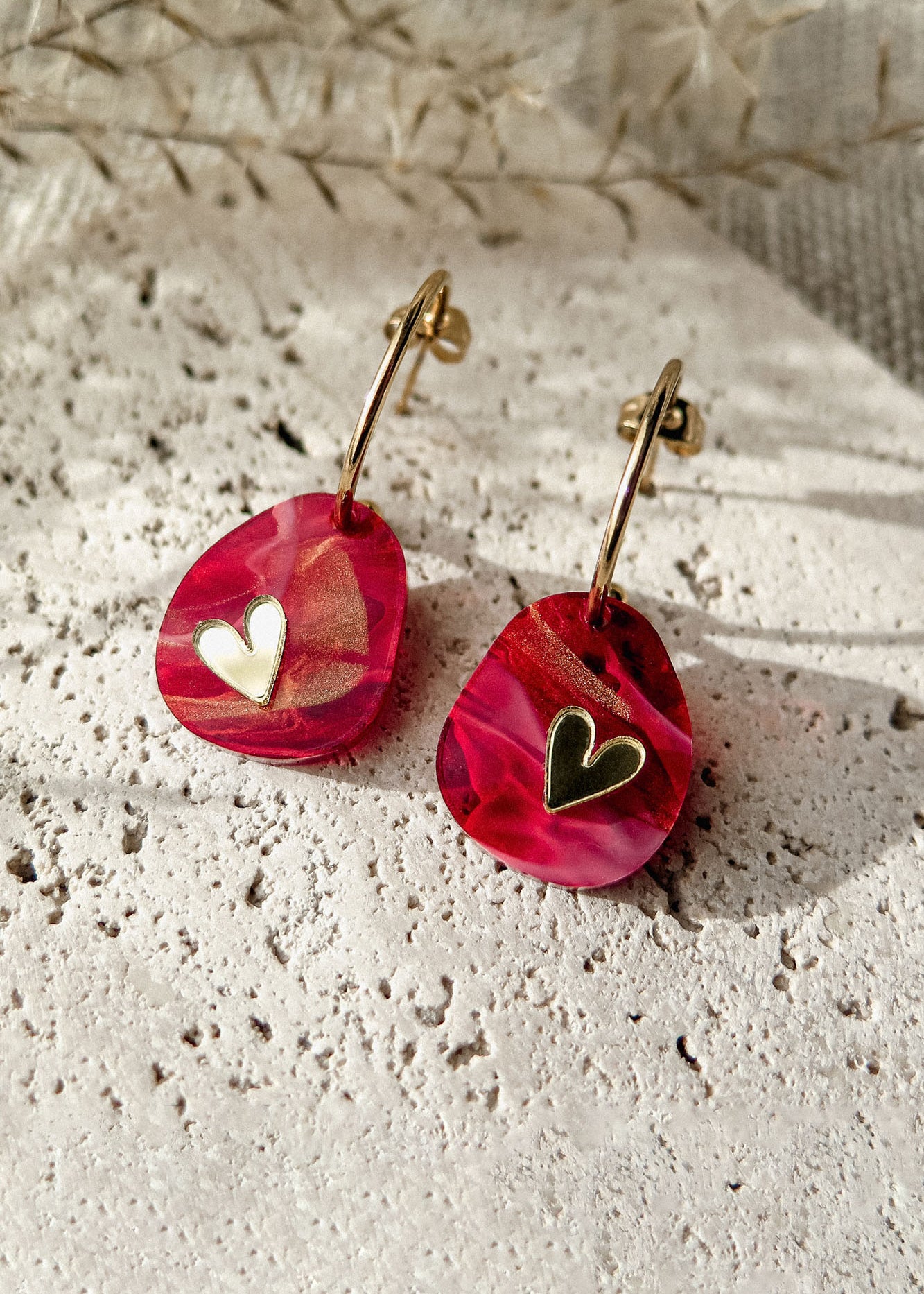 A pair of gold hoop Valentine Pebble Earrings with marbled pink resin pendants, each featuring a small gold heart detail, resting on a textured white stone surface—a romantic accessory perfect for love-inspired style.