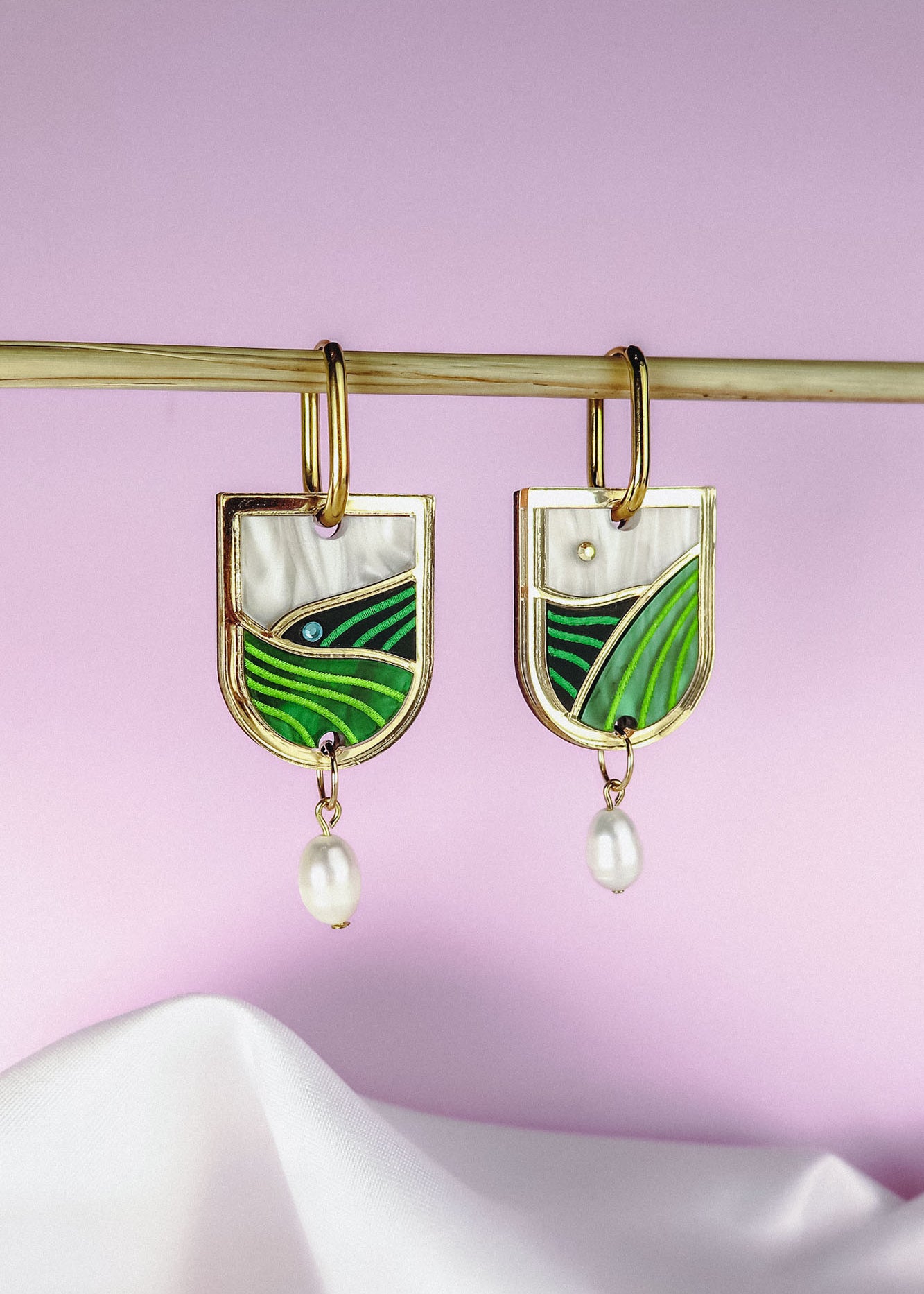 Gold Vineyard Inspired Art Nouveau Dangle Earrings with green and white enamel, an abstract landscape design, and small pearl drops, hang from a horizontal rod against a soft pink and white background.