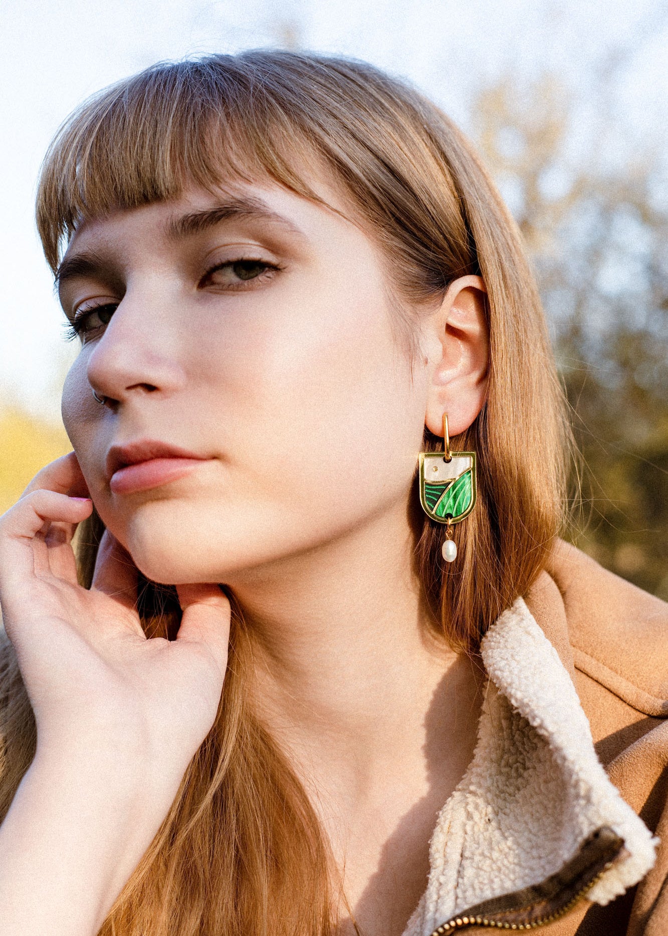 A young woman with light brown hair and bangs gazes calmly at the camera. She wears a tan fleece jacket and green and gold Art Nouveau earrings with a pearl drop. Her hand rests gently on her face, trees blurred in the background.