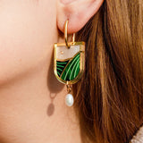 Close-up of a woman's ear wearing handcrafted acrylic dangle earrings with a geometric green and white design, featuring a small pearl drop. The background shows light brown hair and skin.