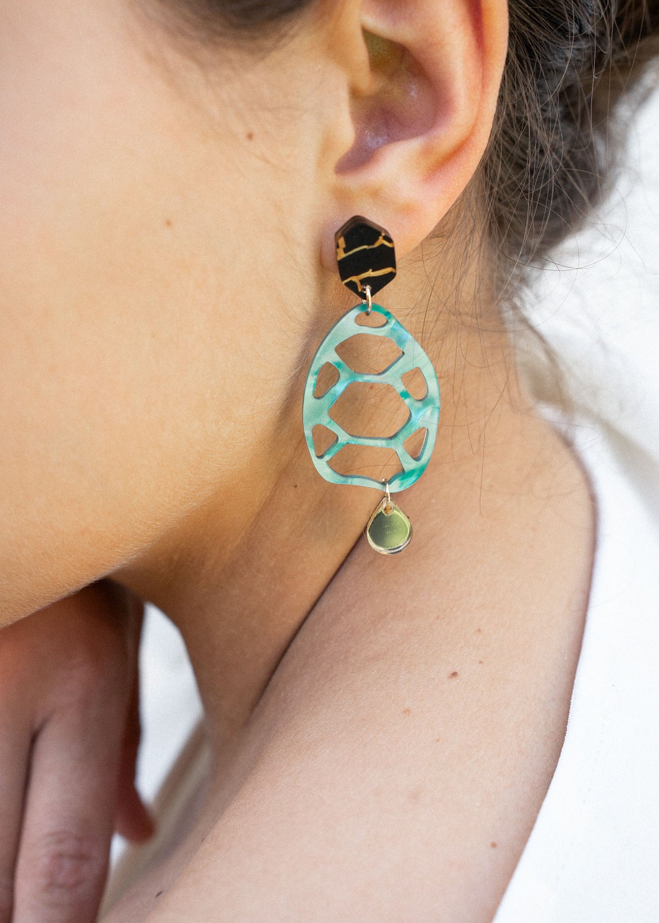 A close-up of a person’s ear wearing a statement, sustainable earring with a black geometric stud, a large mint green oval with cutouts, and a small gold disc at the bottom—showcasing zero-waste beauty. The person has light skin and dark brown hair.