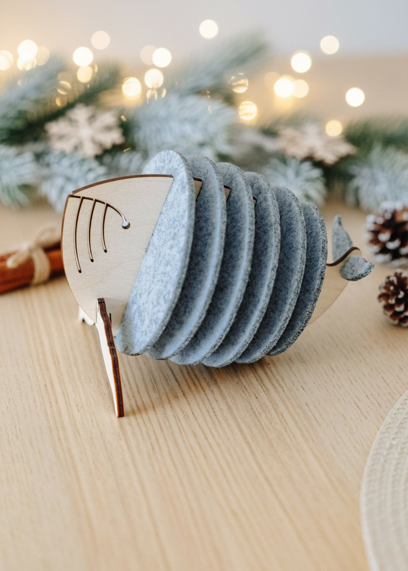 A set of cute coasters held in a wooden holder shaped like a whale, placed on a light wooden table with pine cones, fir branches, and warm fairy lights in the background—a charming touch for your home decor.