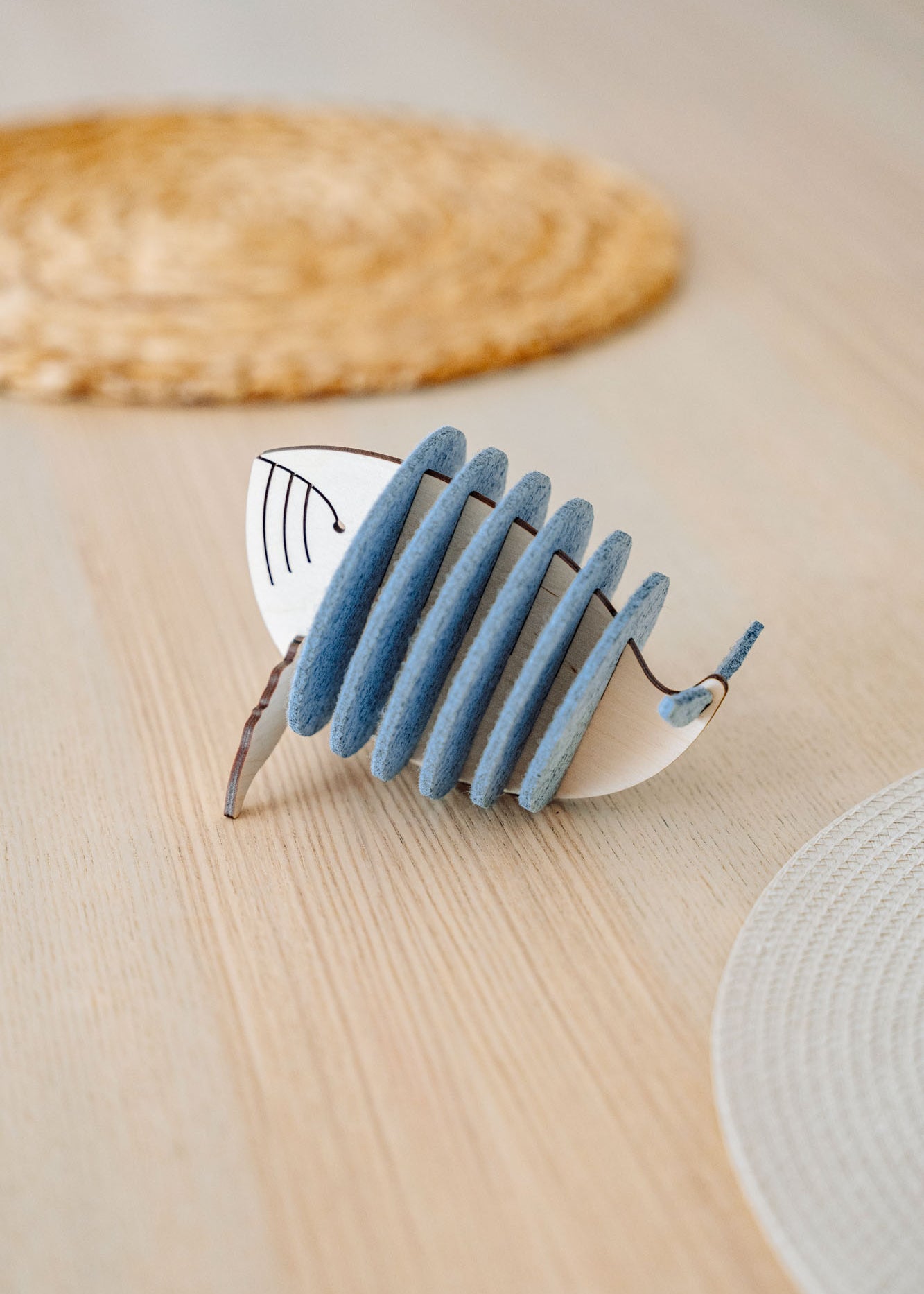 A decorative object resembling a stylized fish, made of alternating blue-grey felt and white pieces, sits on a light wooden table with woven placemats in the background, adding home charm and protecting surfaces like unique Whale Coasters.